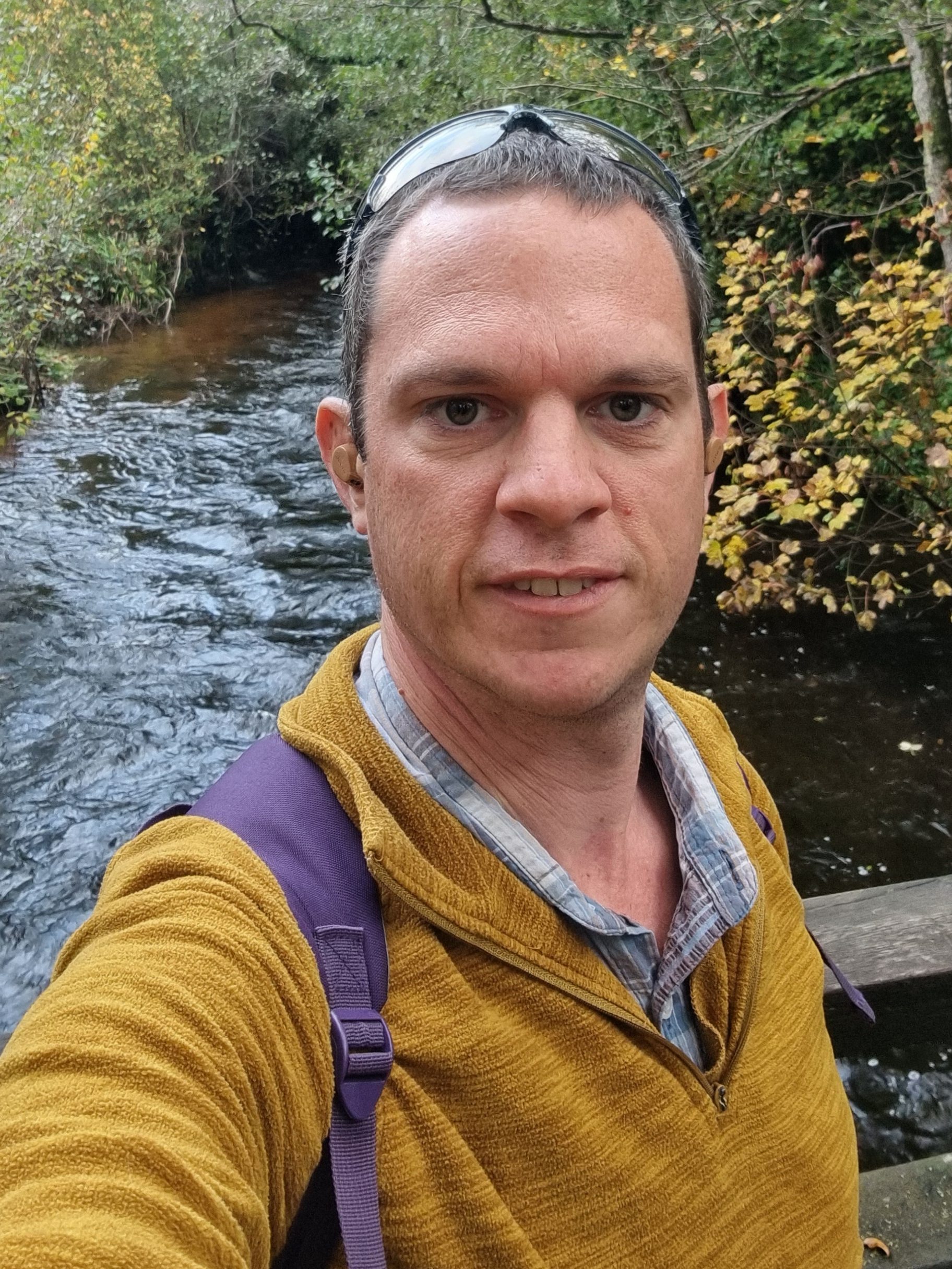 Simon Bates wearing a yellow jumper standing on a bridge over a stream.